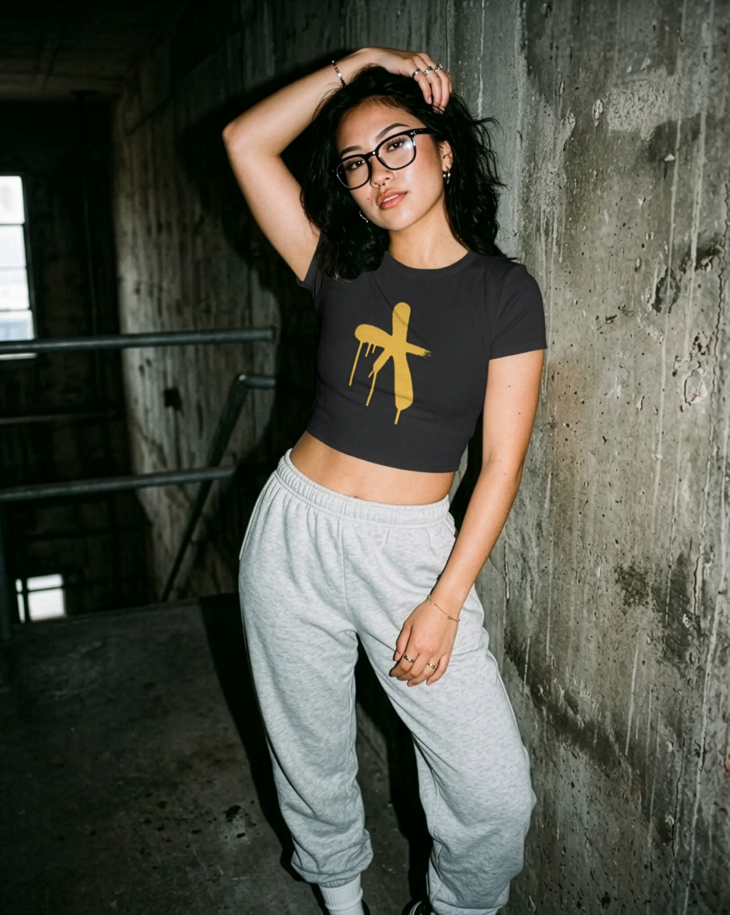 Attractive girl wearing a black crop top with a yellow graphic and grey sweatpants in an industrial rugged setting.