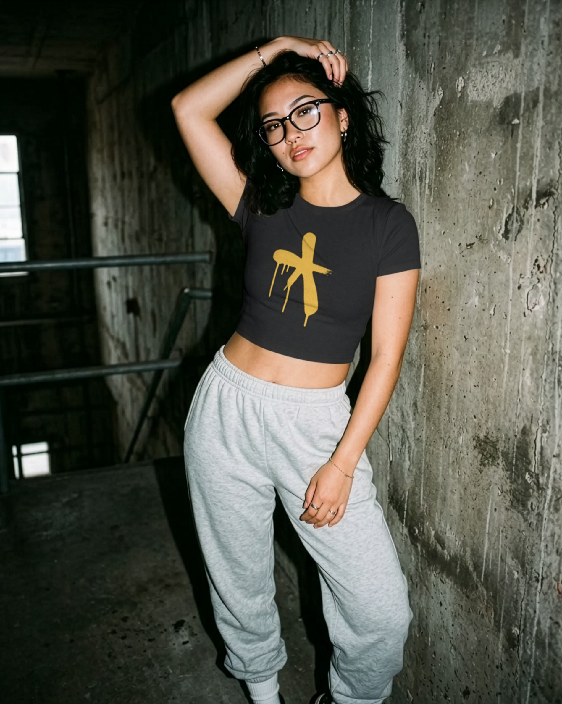 Attractive girl wearing a black crop top with a yellow graphic and grey sweatpants in an industrial rugged setting.